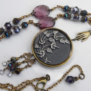 May include: A necklace featuring a central round black button with a silver floral relief, suspended from a brass chain. The design includes purple faceted beads, clear crystals, and a small brass hand charm, finished with a lobster claw clasp.