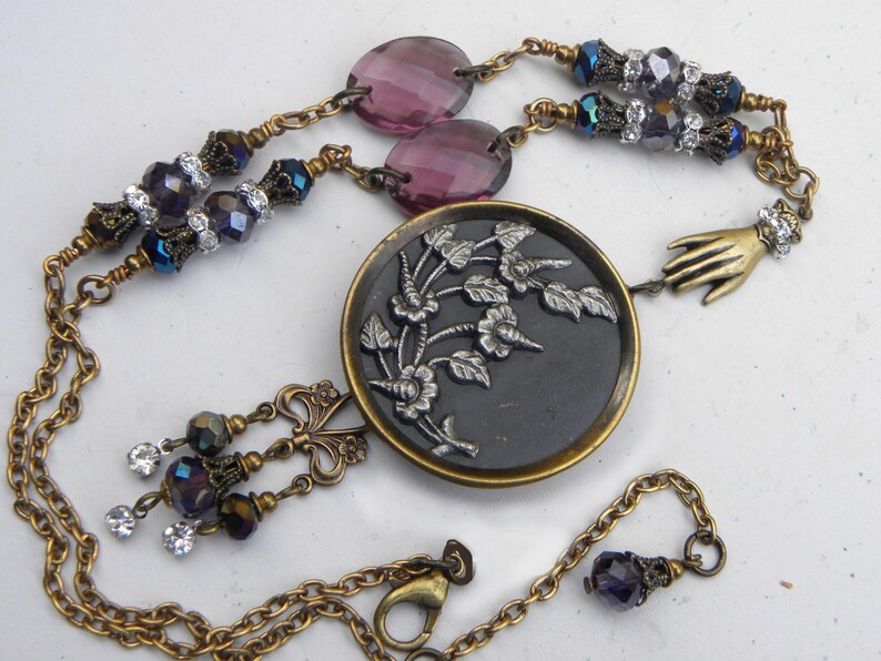 May include: A necklace featuring a central round black button with a silver floral relief, suspended from a brass chain. The design includes purple faceted beads, clear crystals, and a small brass hand charm, finished with a lobster claw clasp.