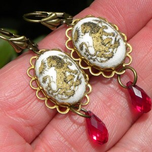 May include: A pair of gold tone earrings with white porcelain cameo pendants featuring a gold-toned design. Each earring has a dangling red crystal drop.