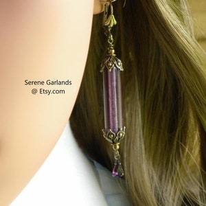 May include: A long, dangling earring with a cylindrical, purple glass tube encased in ornate, antique gold-colored metal. A small, purple teardrop-shaped bead hangs from the bottom. The text "Serene Garlands @ Etsy.com" is visible.