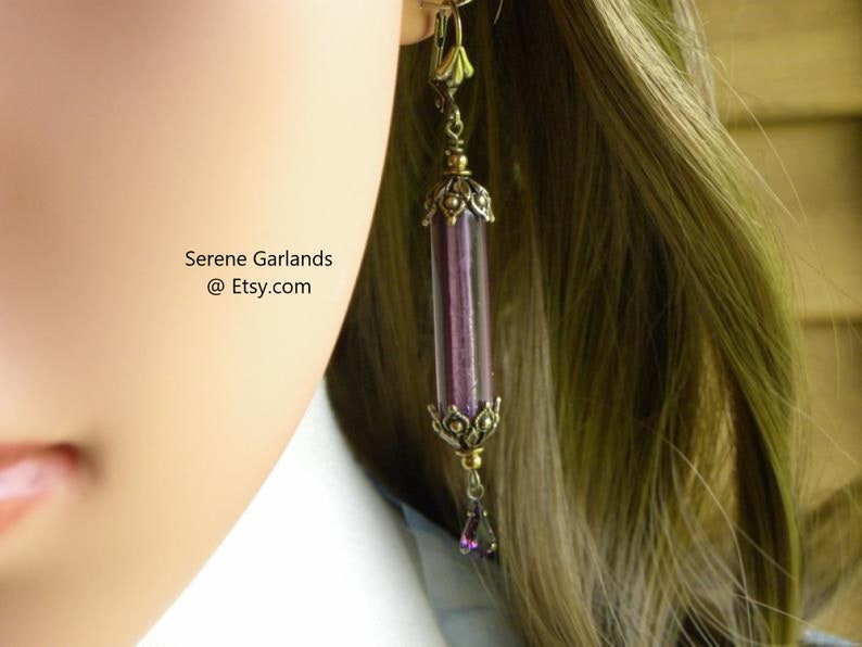 May include: A long, dangling earring with a cylindrical, purple glass tube encased in ornate, antique gold-colored metal. A small, purple teardrop-shaped bead hangs from the bottom. The text "Serene Garlands @ Etsy.com" is visible.