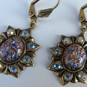 Pode incluir: Um par de brincos de bronze com um design de raios de sol. Cada brinco apresenta um caboch&atilde;o redondo de vidro iridescente rodeado por strass transparentes.