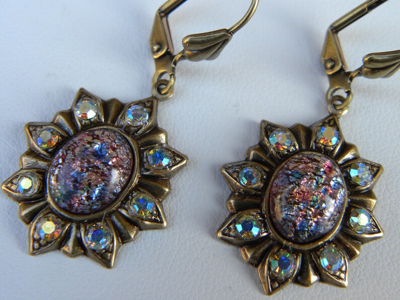 Pode incluir: Um par de brincos de bronze com um design de raios de sol. Cada brinco apresenta um caboch&atilde;o redondo de vidro iridescente rodeado por strass transparentes.