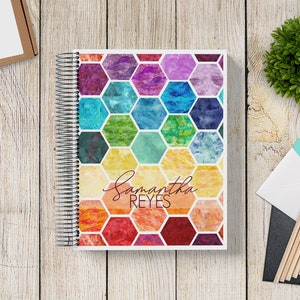 May include: A colorful spiral bound notebook with a honeycomb pattern and the name "Samantha Reyes" written in cursive on the front cover.