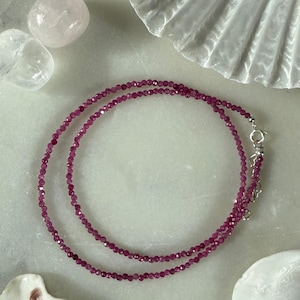 May include: A delicate necklace featuring small, faceted, deep pink gemstones strung together. The necklace is secured with a silver-colored clasp. The background includes decorative elements like a seashell and crystals, enhancing the jewelry's presentation.