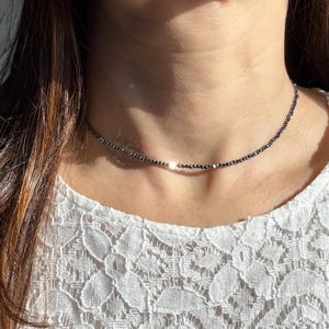 Könnte beinhalten: Eine zarte Choker-Halskette aus kleinen, dunklen Perlen. Die Halskette wird um den Hals getragen und liegt auf einem weißen Spitzentop. Die Perlen scheinen gleichmäßig verteilt zu sein und erzeugen ein subtiles, elegantes Design.