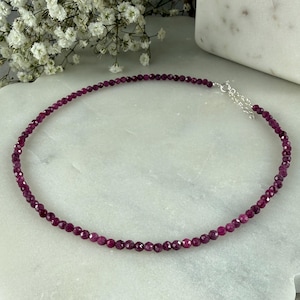 May include: A delicate necklace featuring small, faceted ruby beads. The necklace is a deep, rich red color and has a silver-toned clasp. The beads are strung closely together, creating a continuous line of color. The necklace is displayed on a white marble surface.