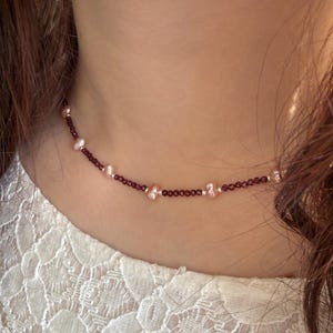 Puede incluir: Un collar delicado con pequeñas cuentas redondas de color rojo oscuro intercaladas con perlas más grandes de color rosa claro. El collar se lleva alrededor del cuello, mostrando su elegante diseño y contraste de color. Es una joya.