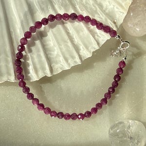 May include: A bracelet with faceted, deep red beads. The beads are strung together, forming a complete circle. The bracelet is secured with a silver-coloured clasp. The jewellery is displayed on a white, textured surface.