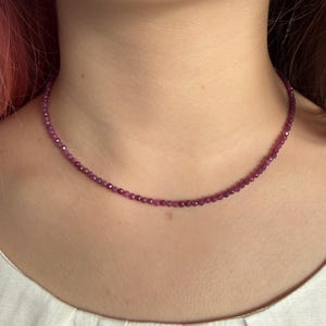 May include: A fine necklace featuring small, round, deep pink beads. The necklace is worn around the neck, highlighting the vibrant colour of the gemstones. The beads are strung together, creating a simple yet elegant piece of jewellery.