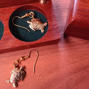 May include: A pair of gold-toned turtle earrings with hook-style backings. One earring is placed inside a circular cutout of a dark green lined wooden box, while the other earring rests on the wooden surface next to the open box.