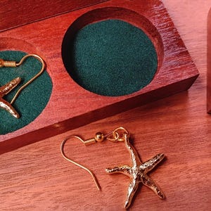 May include: A pair of gold-toned starfish earrings with hook-style backings. One earring is placed inside a circular cutout of a polished wooden box lined with dark green fabric, while the other earring rests on the wooden surface nearby.