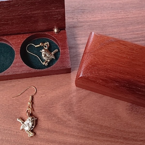 May include: A pair of gold-toned bird-shaped earrings with hook-style backings. One earring is placed inside a small, rectangular, dark reddish-brown wooden box with two circular, green-lined compartments, while the other rests outside.