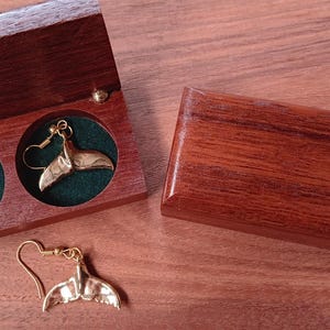 May include: A pair of gold-toned whale tail earrings with hook backings. One earring is placed inside a rectangular wooden box with a dark green velvet interior, while the other earring rests outside the box. A second closed wooden box is also shown.