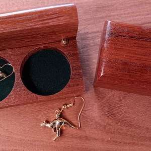 May include: A pair of gold-toned kangaroo-shaped drop earrings with hook-style backings. One earring is placed inside a small, rectangular wooden box with a dark green velvet-lined interior, while the other earring rests outside the box.
