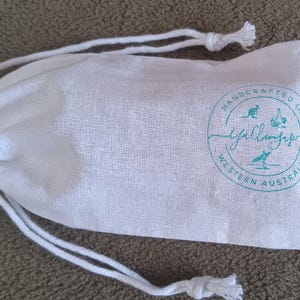 May include: A white fabric drawstring pouch featuring a circular teal logo. The logo includes the text "HANDCRAFTED IN WESTERN AUSTRALIA" surrounding a central script word, along with small icons of a kangaroo, a garlic bulb, and a person surfing.
