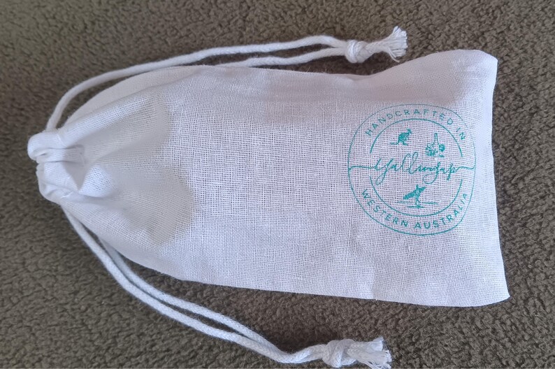 May include: A white fabric drawstring pouch featuring a circular teal logo. The logo includes the text "HANDCRAFTED IN WESTERN AUSTRALIA" surrounding a central script word, along with small icons of a kangaroo, a garlic bulb, and a person surfing.