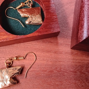 May include: A pair of gold-toned shark head earrings with fishhook wires. One earring is placed inside a circular cutout of a dark reddish-brown wooden box, while the other earring rests outside the box on a matching wooden surface.