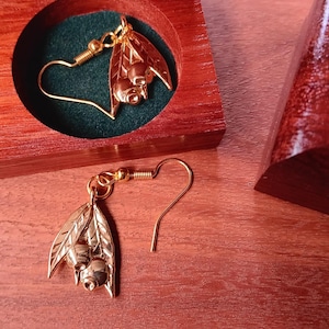 May include: A pair of gold-toned metal earrings featuring a botanical design of two small berries nestled between two leaves. One earring is placed inside a circular cutout of a wooden box with a dark green velvet lining, while the other rests below.