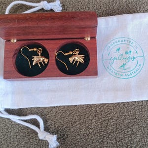 May include: A pair of gold-toned bee-shaped drop earrings with hook backings, presented inside a small rectangular dark wood box with a hinged lid. The box sits on a white fabric drawstring pouch featuring a teal circular logo stamp.
