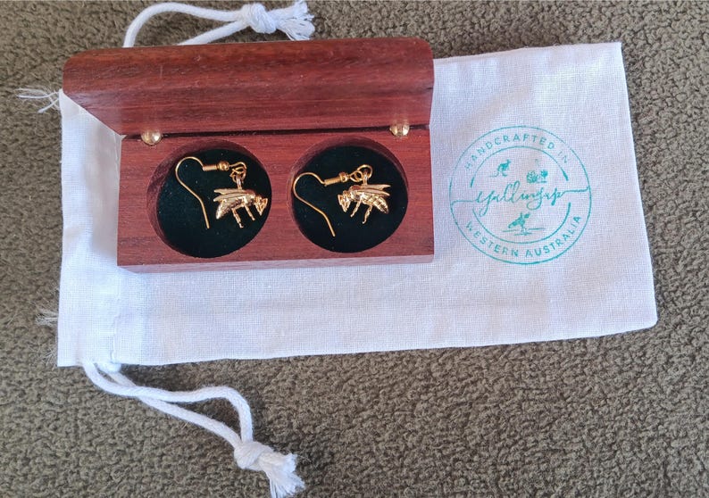 May include: A pair of gold-toned bee-shaped drop earrings with hook backings, presented inside a small rectangular dark wood box with a hinged lid. The box sits on a white fabric drawstring pouch featuring a teal circular logo stamp.