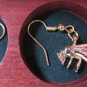 May include: A pair of gold-toned bee-shaped dangle earrings with hook-style ear wires. Each earring features a detailed metallic bee charm suspended from a gold-colored wire, presented within the circular cutouts of a dark wooden display case.