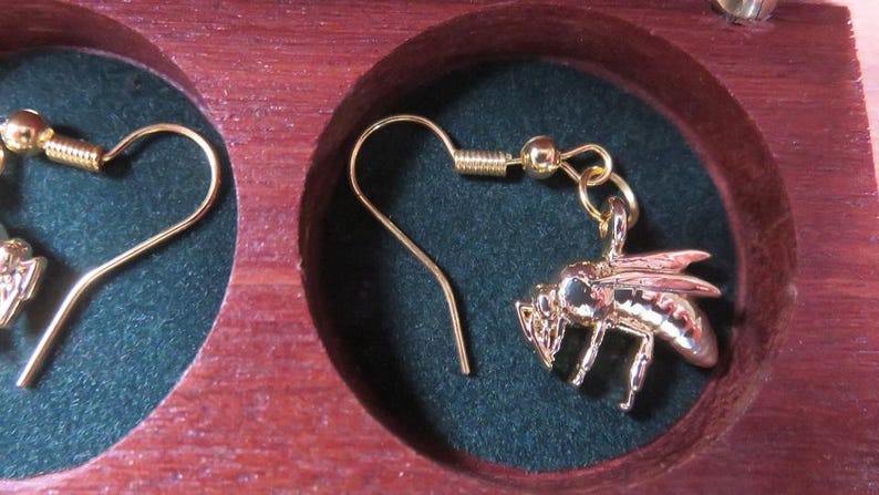 May include: A pair of gold-toned bee-shaped dangle earrings with hook-style ear wires. Each earring features a detailed metallic bee charm suspended from a gold-colored wire, presented within the circular cutouts of a dark wooden display case.