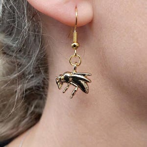 May include: A gold-toned metal earring featuring a detailed bee charm suspended from a standard fishhook wire. The bee charm has a polished, reflective surface with visible wings, legs, and body segments, hanging vertically from a small circular jump ring.