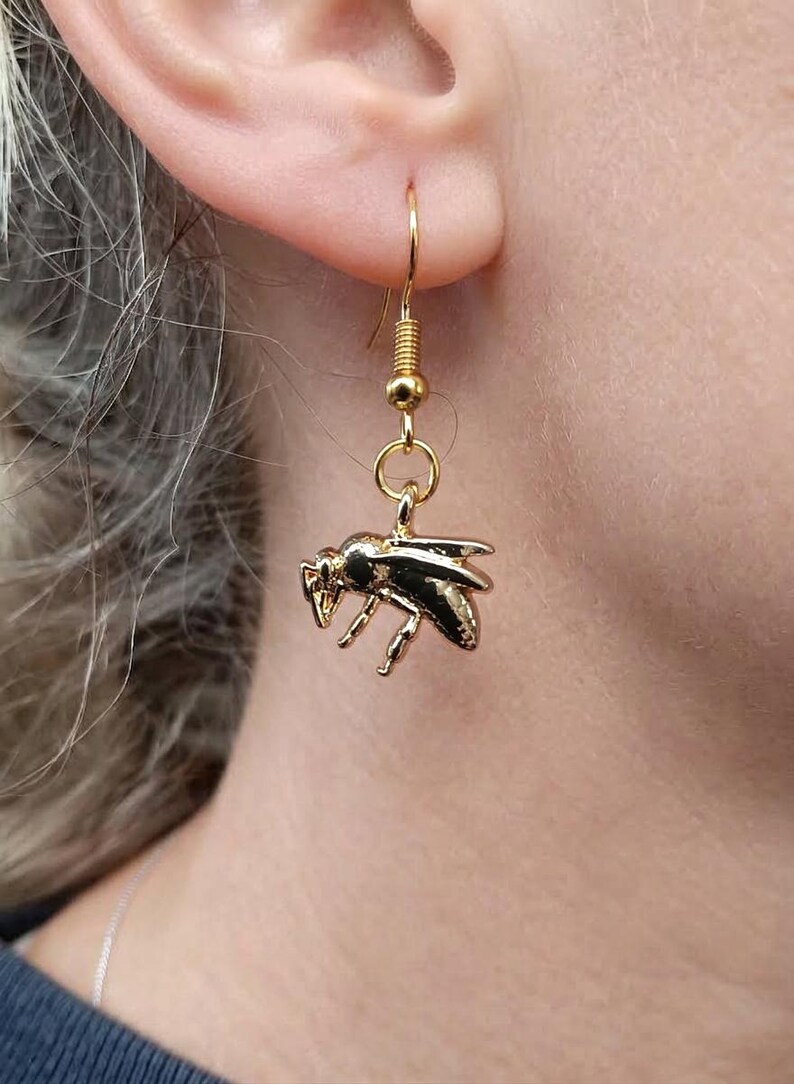 May include: A gold-toned metal earring featuring a detailed bee charm suspended from a standard fishhook wire. The bee charm has a polished, reflective surface with visible wings, legs, and body segments, hanging vertically from a small circular jump ring.