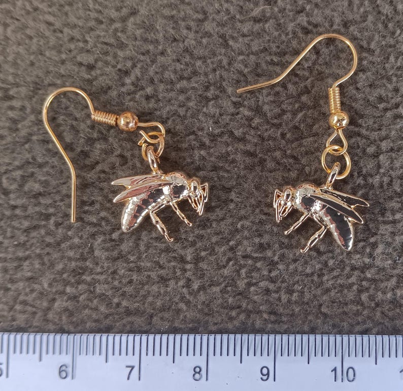 May include: A pair of gold-toned bee-shaped dangle earrings with hook-style wires. The bee charms feature detailed wings and body segments. A ruler is visible at the bottom, showing the bee charms measure approximately 0.75 inches in width.