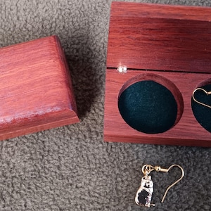 May include: A pair of gold-toned cat-shaped dangle earrings with hook backings. One earring is placed inside a rectangular wooden box with a dark green velvet interior, while the other earring rests outside the box next to the open lid.