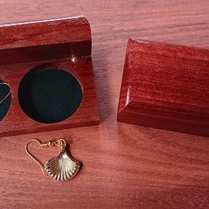 May include: A pair of gold-toned shell-shaped earrings with hook backings. One earring is placed inside a dark reddish-brown wooden box with two circular velvet-lined compartments, while the other earring rests outside the open box.