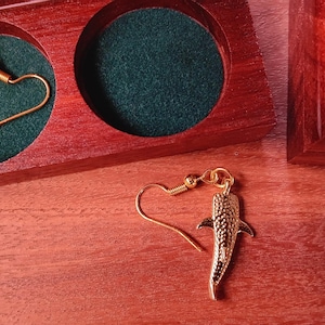 May include: A pair of gold-toned whale shark earrings with textured skin details and fishhook-style ear wires. One earring is placed inside a circular cutout of a dark red wooden box with a green velvet lining, while the other rests outside.