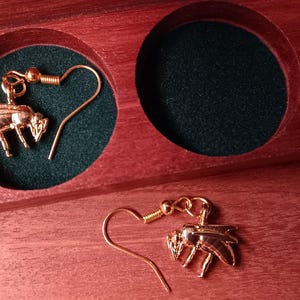 May include: A pair of gold-toned bee-shaped drop earrings with hook-style backings. One earring is placed inside a circular cutout of a dark green velvet-lined wooden box, while the other earring rests on the wooden surface next to the box.
