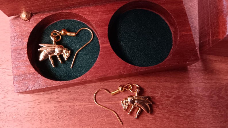 May include: A pair of gold-toned bee-shaped drop earrings with hook-style backings. One earring is placed inside a circular cutout of a dark green velvet-lined wooden box, while the other earring rests on the wooden surface next to the box.