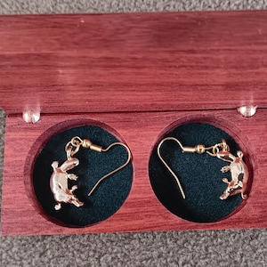 May include: A pair of gold-toned platypus-shaped drop earrings with hook-style backings, displayed inside a rectangular wooden box featuring a dark red finish and two circular compartments lined with dark green velvet fabric.
