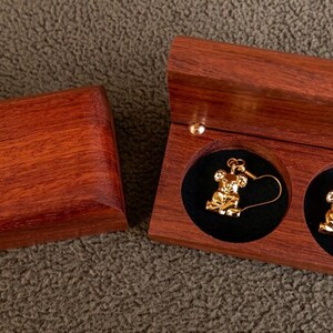 May include: A pair of gold-toned koala-shaped dangle earrings with hook backings, presented inside a rectangular wooden box with a hinged lid and two circular black velvet-lined compartments for secure storage of the jewelry pieces.
