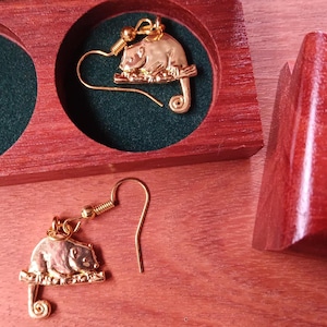 May include: A pair of gold-toned metal earrings featuring a small animal perched on a branch with a curled tail. One earring is placed inside a circular cutout of a dark red wooden box, while the other rests on the surface next to the box.