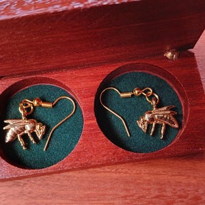 May include: A pair of gold-toned bee-shaped dangle earrings with hook-style backings, resting inside a rectangular wooden box featuring two circular compartments lined with dark green fabric and a hinged lid that is open.