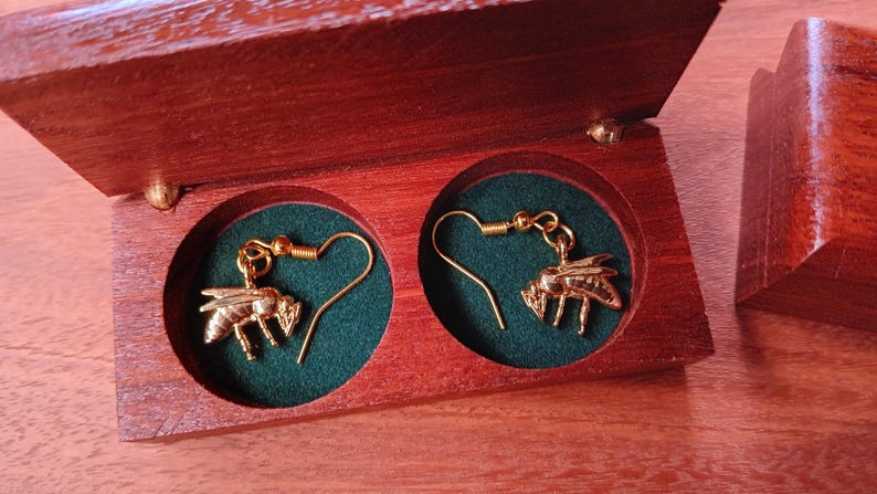May include: A pair of gold-toned bee-shaped dangle earrings with hook-style backings, resting inside a rectangular wooden box featuring two circular compartments lined with dark green fabric and a hinged lid that is open.