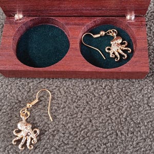 May include: A pair of gold-toned octopus-shaped drop earrings with hook-style backings. One earring is placed inside a circular cutout of a dark brown wooden box with a green velvet lining, while the other earring rests outside the box.