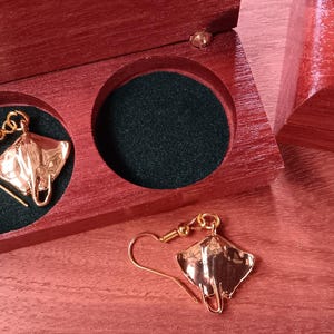 May include: A pair of gold-toned stingray-shaped earrings with hook-style backings. One earring is placed inside a circular cutout of a polished reddish-brown wooden box, while the other earring rests on the surface next to the open box.