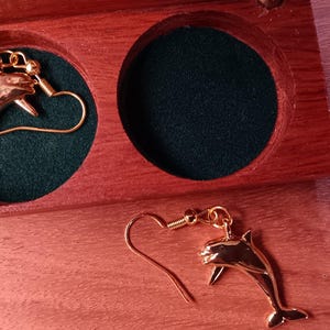 May include: A pair of gold-toned dolphin earrings with hook-style backings. One earring is placed inside a circular cutout of a polished wooden box lined with dark green fabric, while the other earring rests on the wooden surface beside the box.