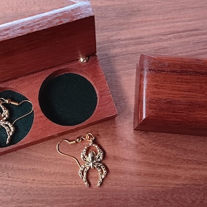 May include: A pair of gold-toned spider-shaped earrings with hook backings. One earring is placed inside a rectangular wooden box with a dark green velvet interior, while the other earring rests outside the box next to a closed wooden box.