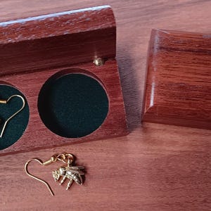 May include: A pair of gold-toned bee-shaped dangle earrings with hook-style backings. One earring is placed inside a circular cutout of a dark brown wooden box with a green velvet lining, while the other earring rests outside the box.