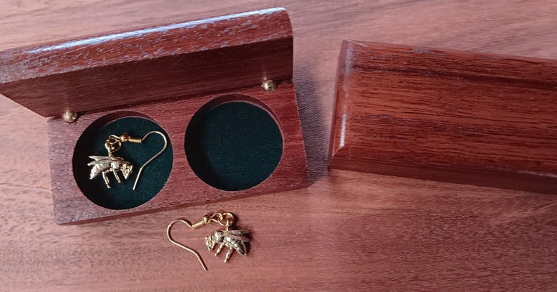 May include: A pair of gold-toned bee-shaped dangle earrings with hook-style backings. One earring is placed inside a circular cutout of a dark brown wooden box with a green velvet lining, while the other earring rests outside the box.