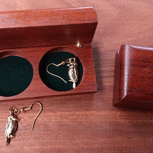 May include: A pair of gold-toned bird-shaped dangle earrings featuring hook-style closures. One earring is placed inside a small, rectangular wooden box with two circular, dark green velvet-lined compartments, while the other rests outside.