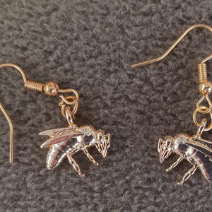 May include: A pair of gold-toned metal drop earrings featuring a detailed bee charm. Each earring consists of a curved ear wire hook connected to a small jump ring, which suspends a polished bee pendant with visible wing and body segments.
