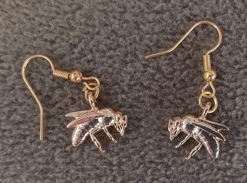 May include: A pair of gold-toned metal drop earrings featuring a detailed bee charm. Each earring consists of a curved ear wire hook connected to a small jump ring, which suspends a polished bee pendant with visible wing and body segments.