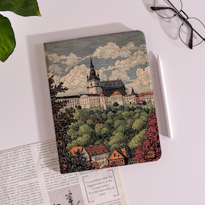 May include: A rectangular notebook featuring a detailed illustration of a European-style town with a prominent church spire, lush green trees, and orange-roofed houses under a cloudy sky, accompanied by a white stylus and black-rimmed glasses.
